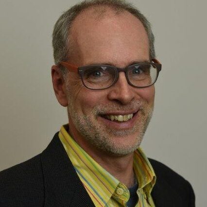 Mark R. (Accelerate Participant with the IMPACT Center). white male with red and brown glasses wearing a block suit with a yellow shirt, looking at camera and smiling.